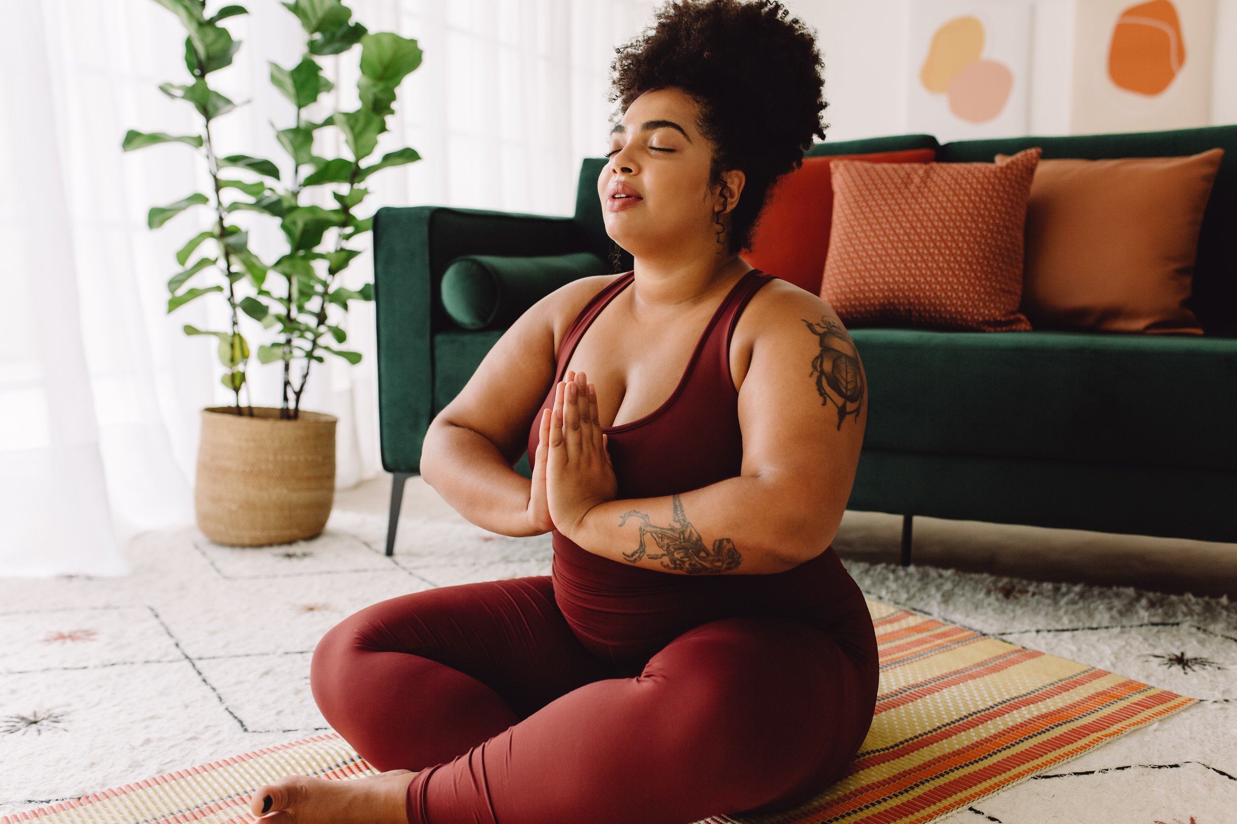 A woman practicing yoga in a living room, sitting cross-legged on a mat with her hands in prayer position, eyes closed, and a calm expression.