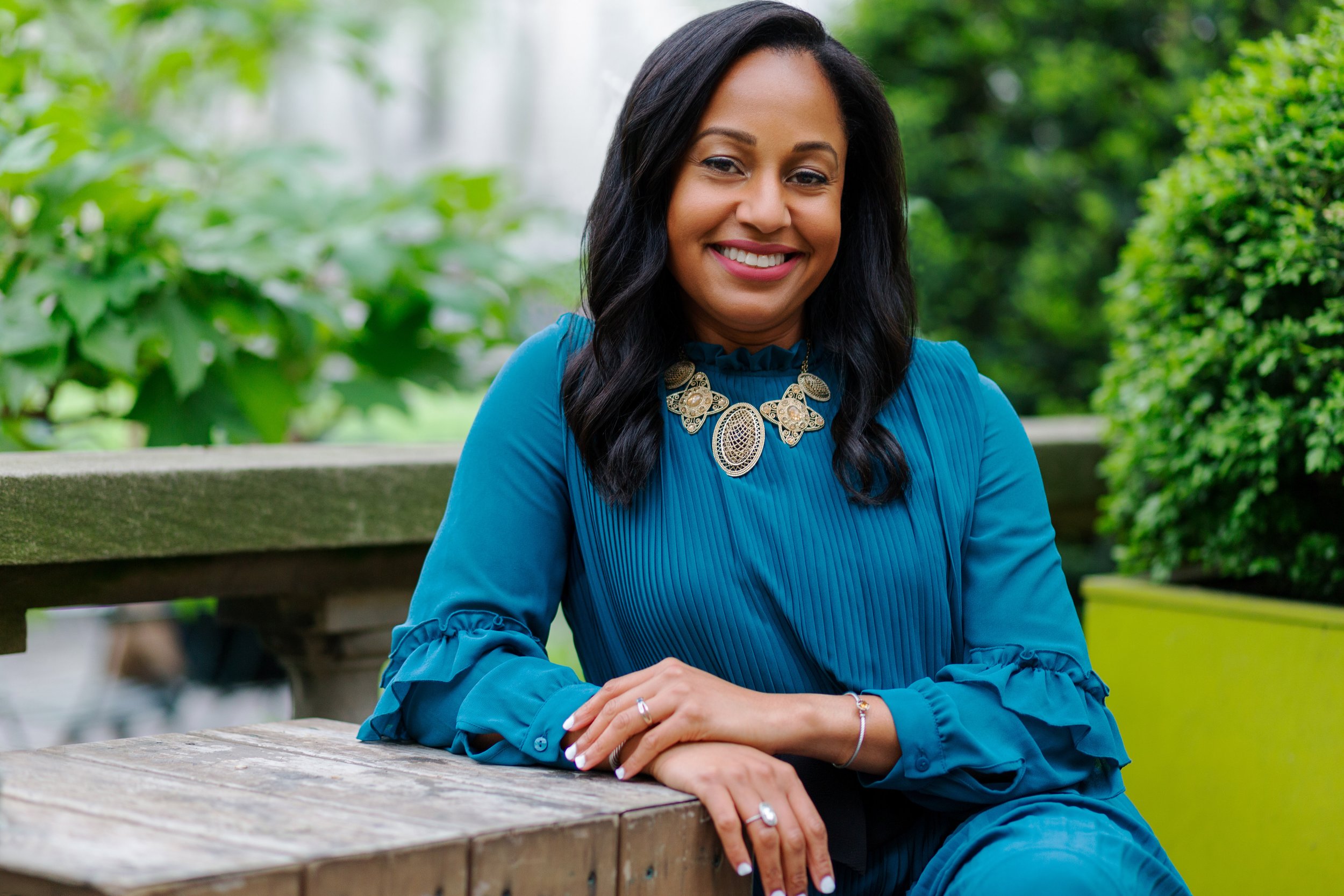 A woman with black hair wearing a blue dress and decorative necklace, smiling and sitting outdoors at a wooden picnic table, with greenery in the background.