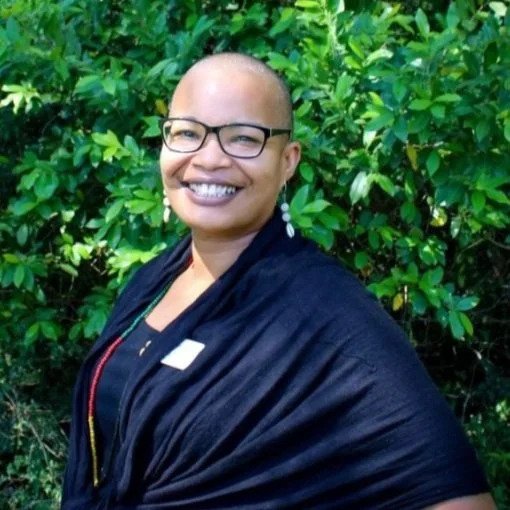Woman with a shaved head, glasses, and earrings smiling outdoors in front of green foliage, wearing a dark shawl over a black top.