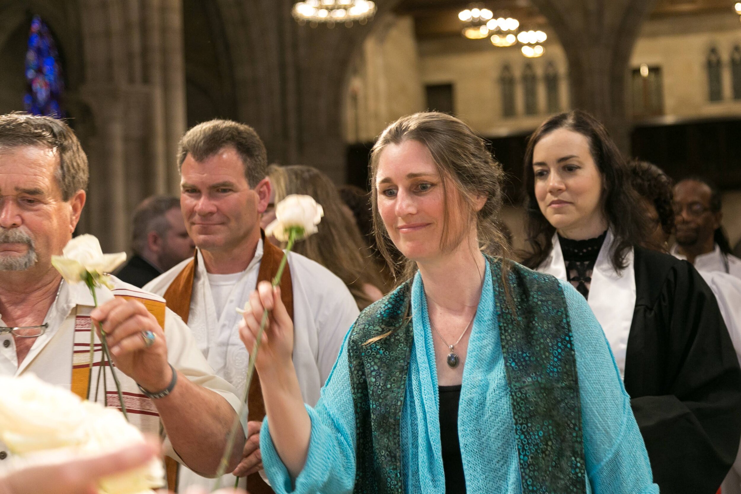 A woman in a turquoise shawl holding a white flower in a church during a religious ceremony, surrounded by people. The church has stained glass windows and ornate architecture.