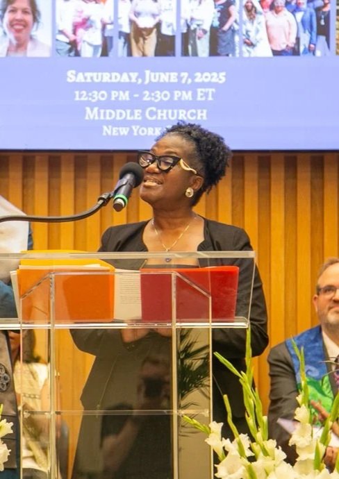 Woman speaking at a podium during a church event, with a large screen behind displaying details of the event and a group photo.