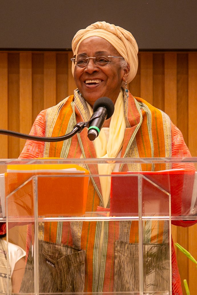 An elderly woman with glasses, wearing a beige headwrap and colorful traditional clothing, speaking at a podium with a microphone in a wood-paneled room.