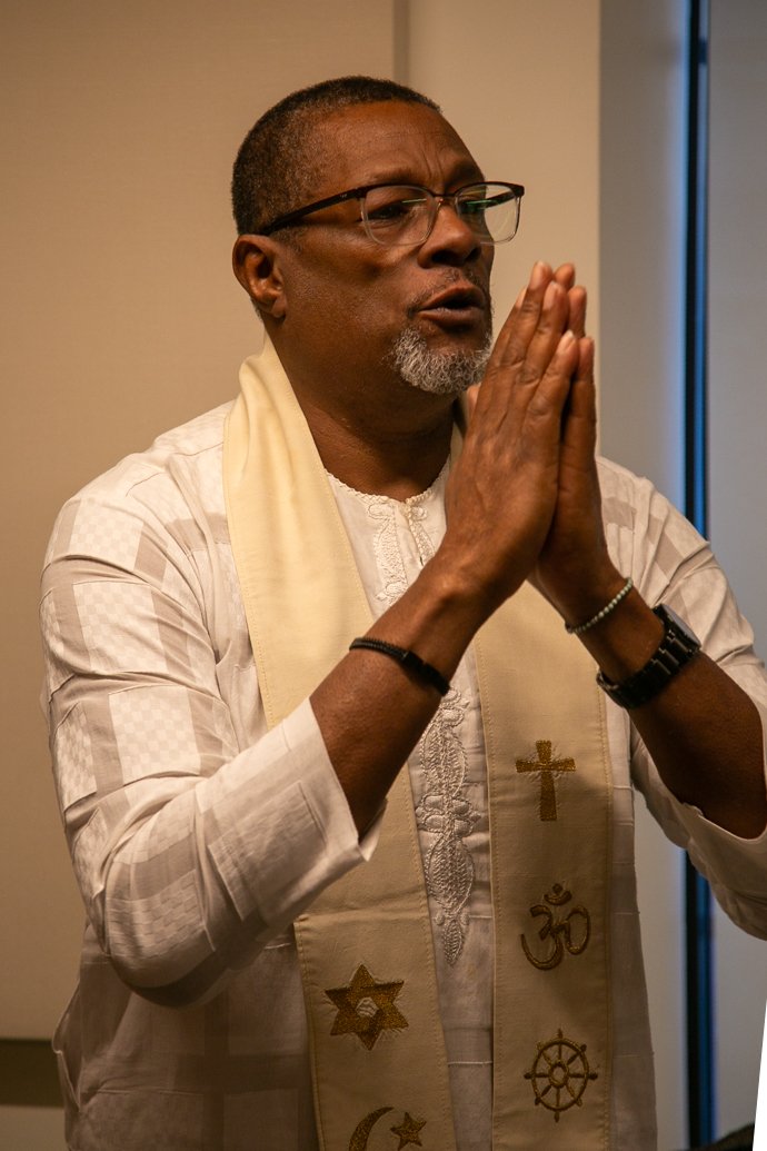 Man praying with hands together, wearing glasses, white traditional attire, and religious stole with symbols.