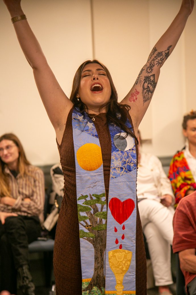 A woman with dark hair and tattoos on her arm is raising her arms and appears to be celebrating or expressing happiness. She is wearing a colorful scarf or garment with various painted symbols, including a sun, a tree, a red heart with drops, and a chalice. Several other people are seated in the background.