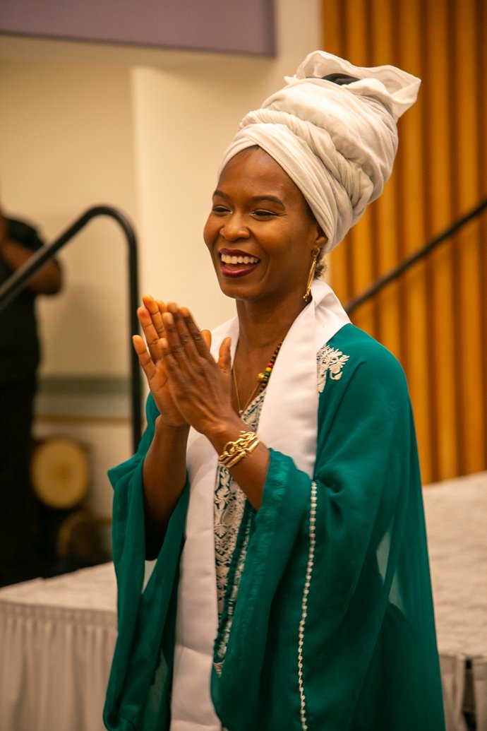 A woman wearing a gray headwrap, teal traditional dress, and gold jewelry is smiling with her hands clasped together.