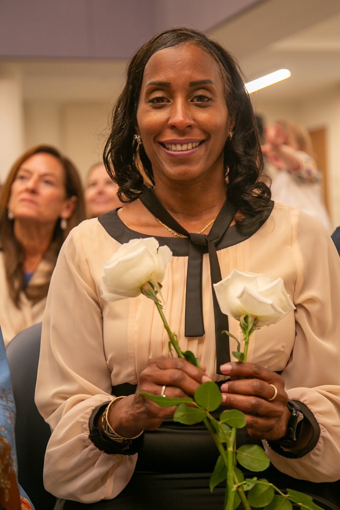A smiling woman holding two white roses at an indoor event, with other attendees in the background.