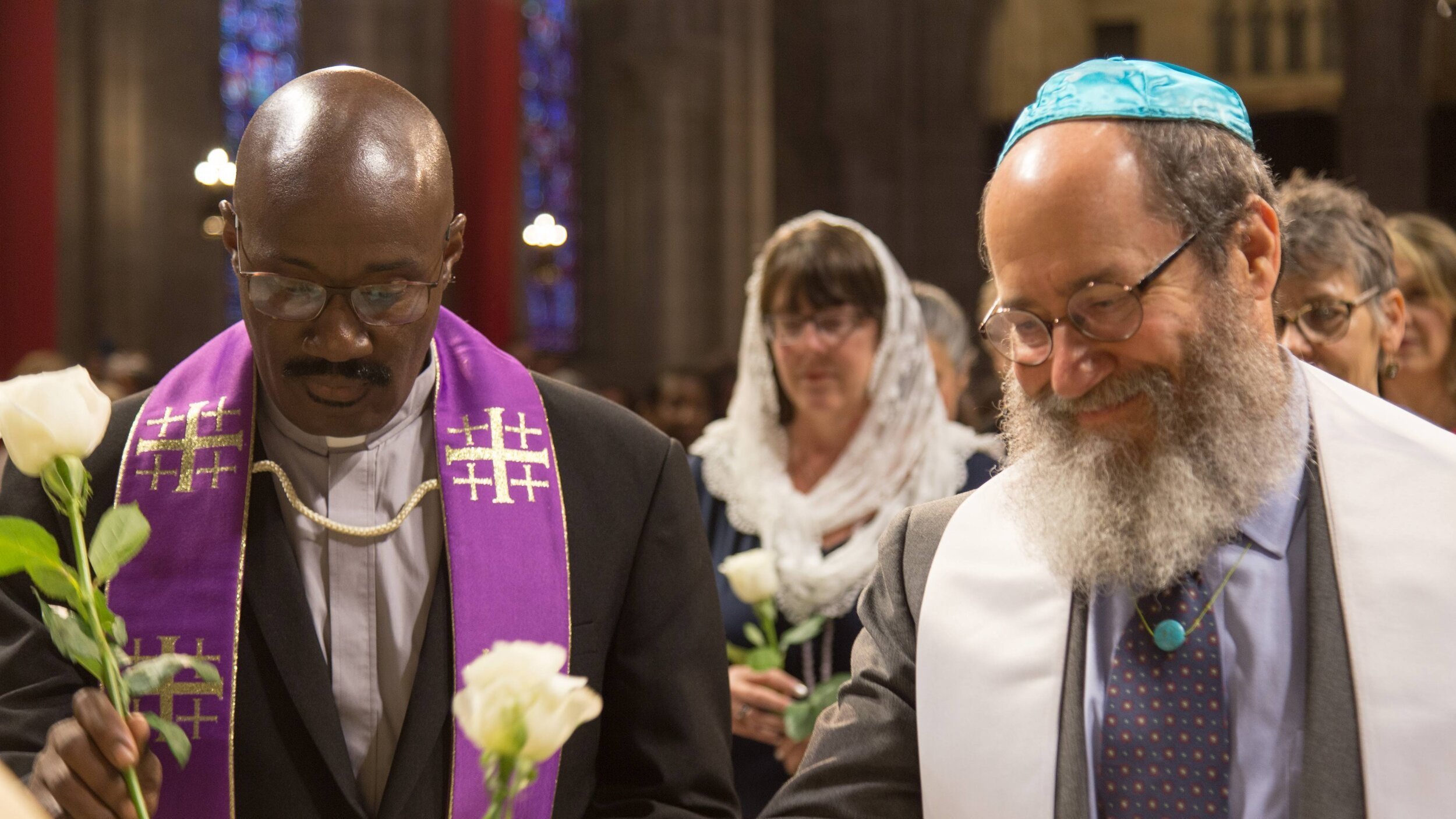 Two men standing in a church, one wearing a purple stole and the other wearing a white tallit, both holding white roses, surrounded by women in traditional Jewish and Christian attire.