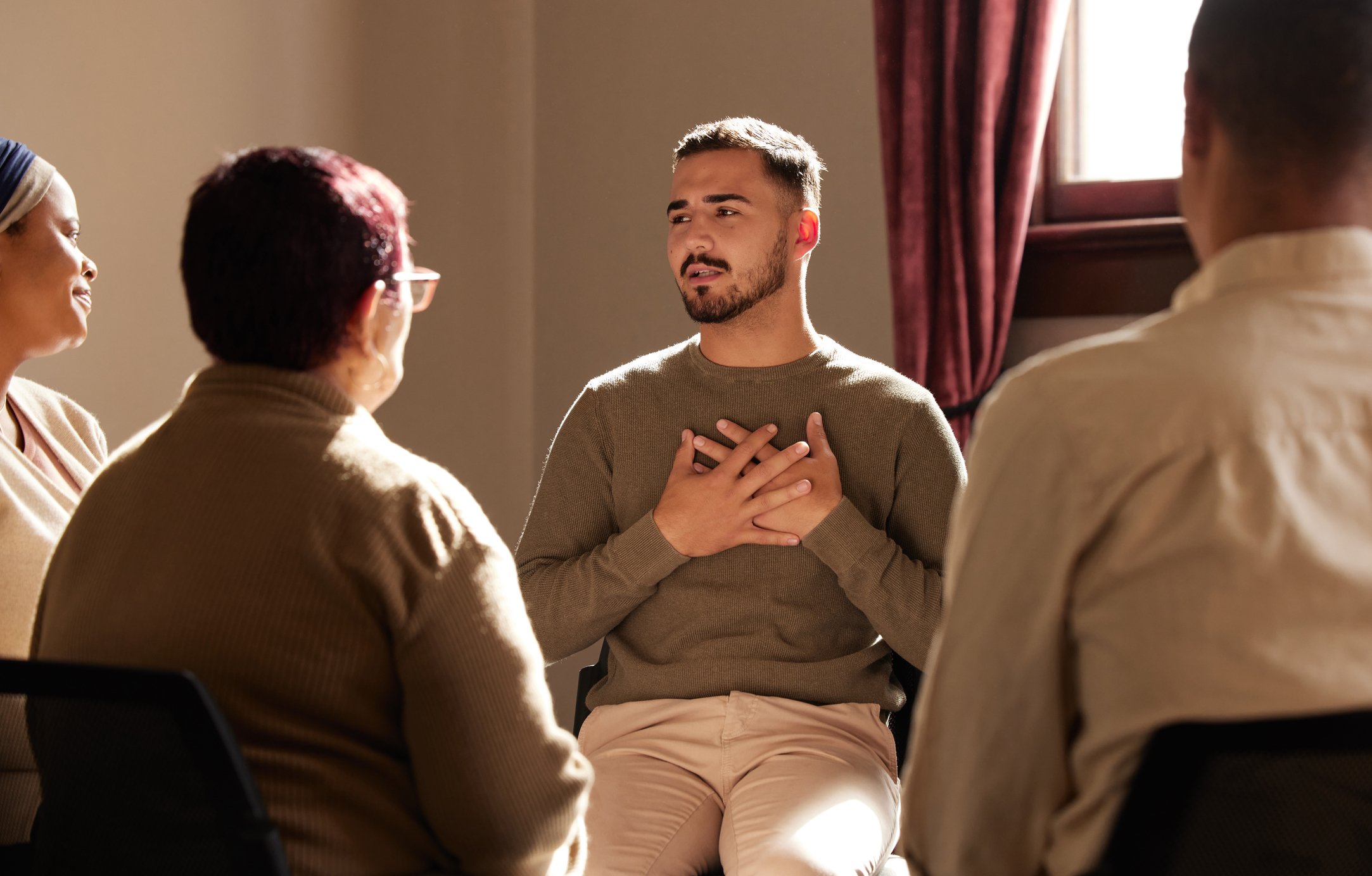 A young man sitting with his hands over his chest, appearing emotional, in a support group session with four women listening attentively in a room with sunlight coming through a window.