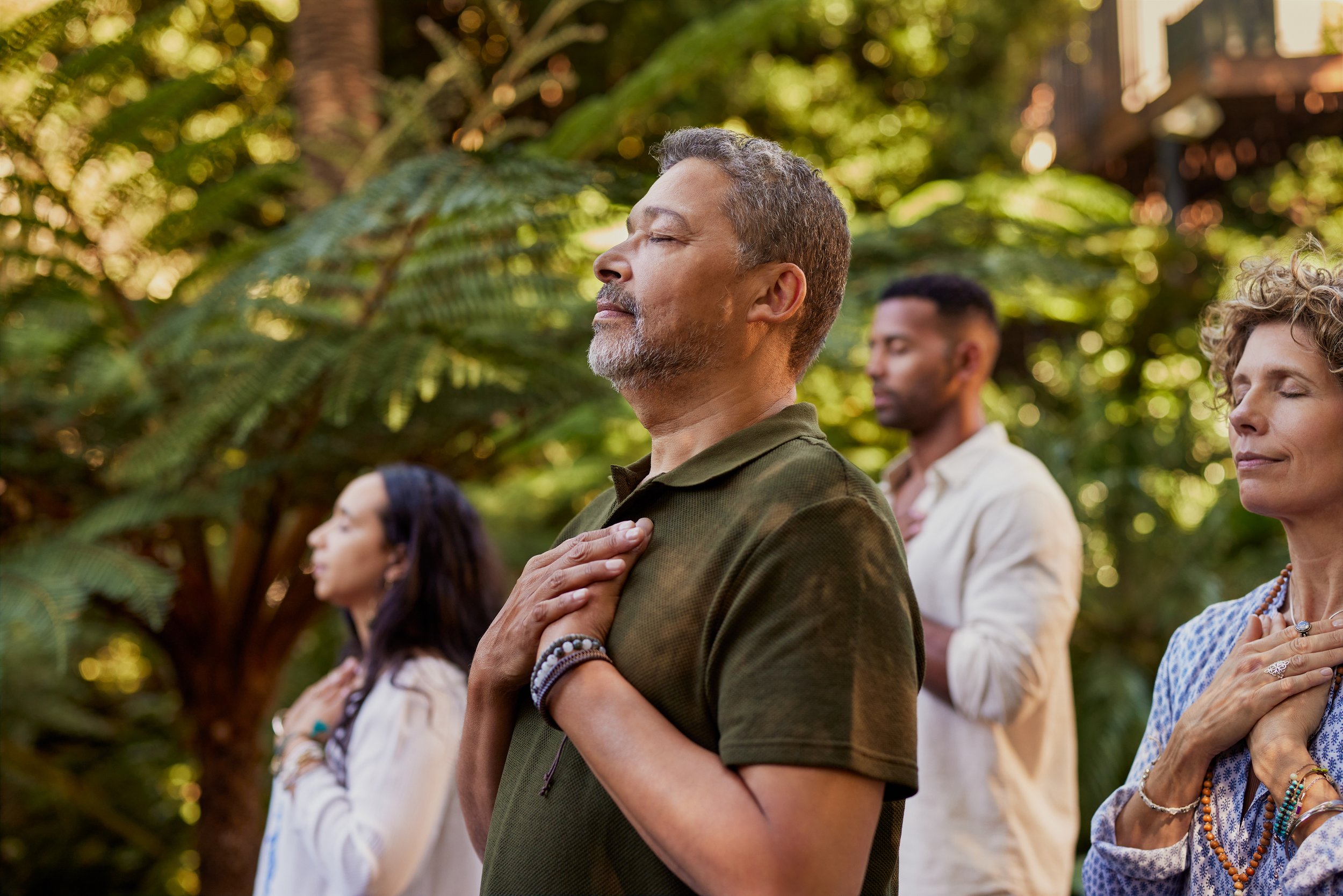 People practicing meditation outdoors with hands on their chests, eyes closed, amid green foliage.