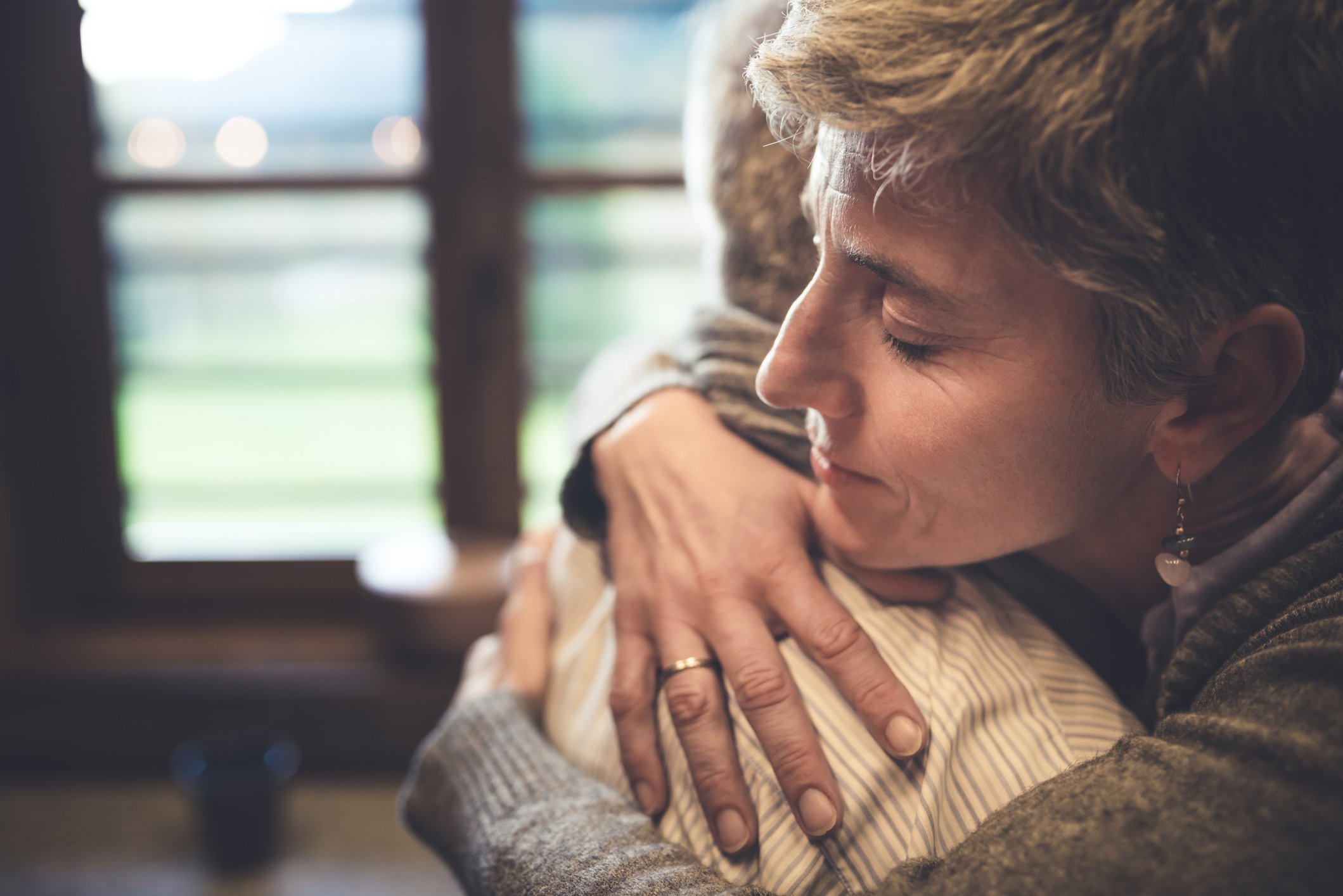 A woman hugging an older person with her eyes closed, inside a warmly lit room with windows in the background.