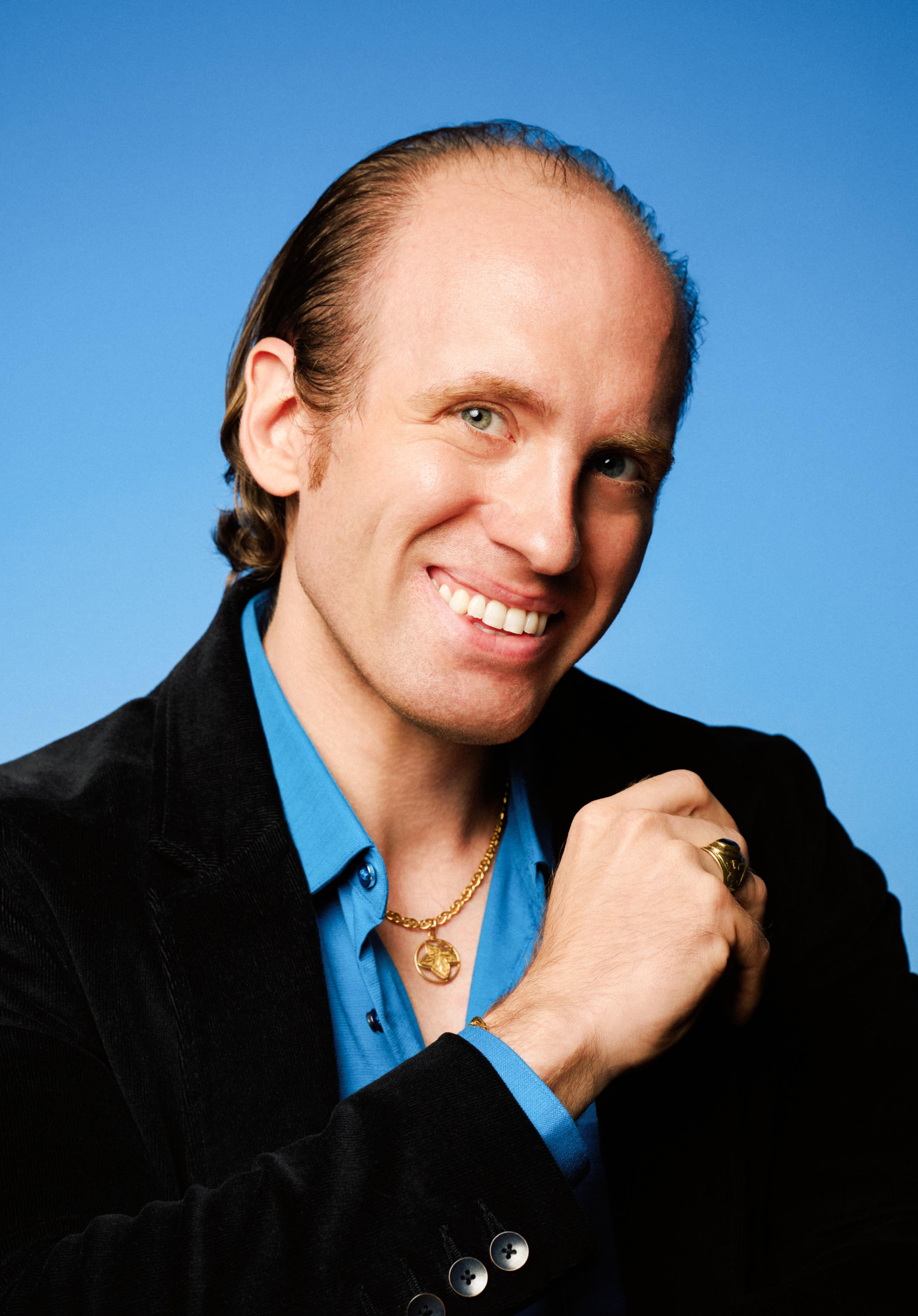 Portrait of a man with light skin, slicked-back brown hair, smiling, wearing a blue shirt, black jacket, gold necklace, and rings, against a blue background.