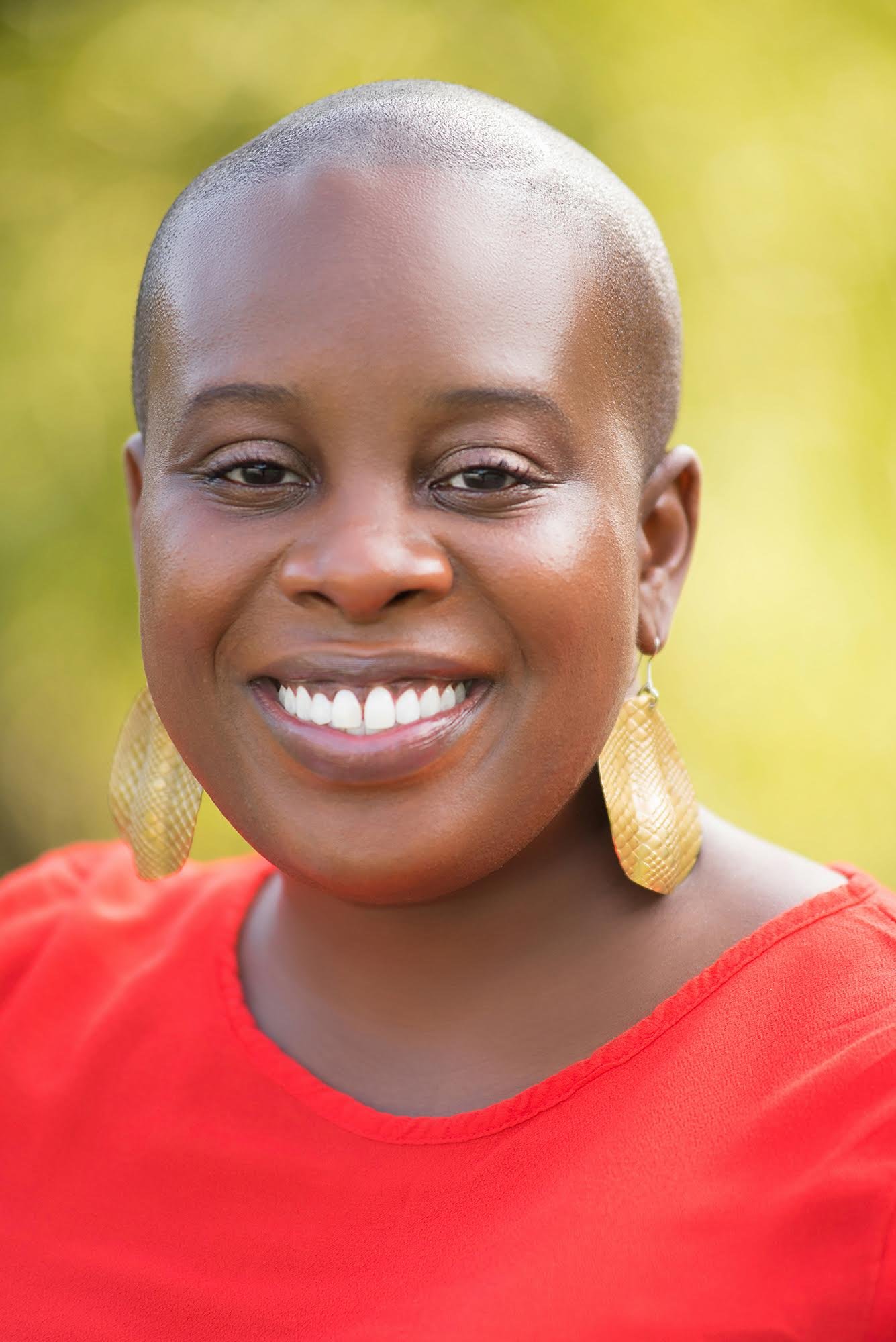 A smiling Black woman with a bald head, wearing gold earrings and a red top, outdoors with a blurred green background.