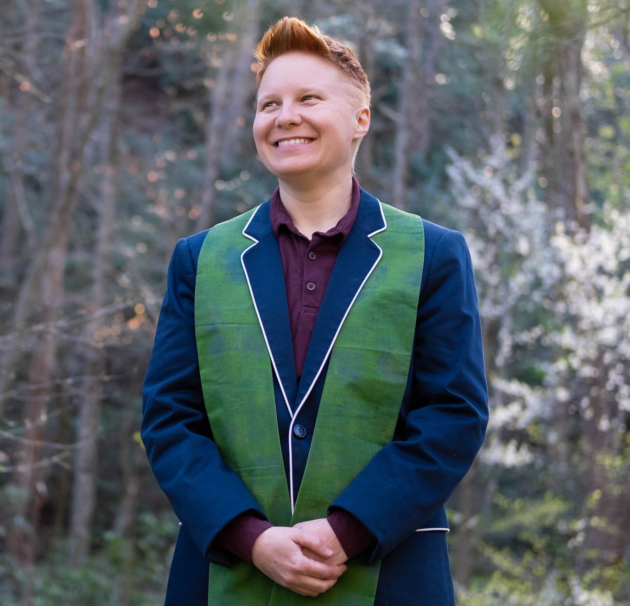 A woman with short red hair smiling outdoors in a forest, wearing a dark blue blazer with green sash and a maroon shirt underneath.