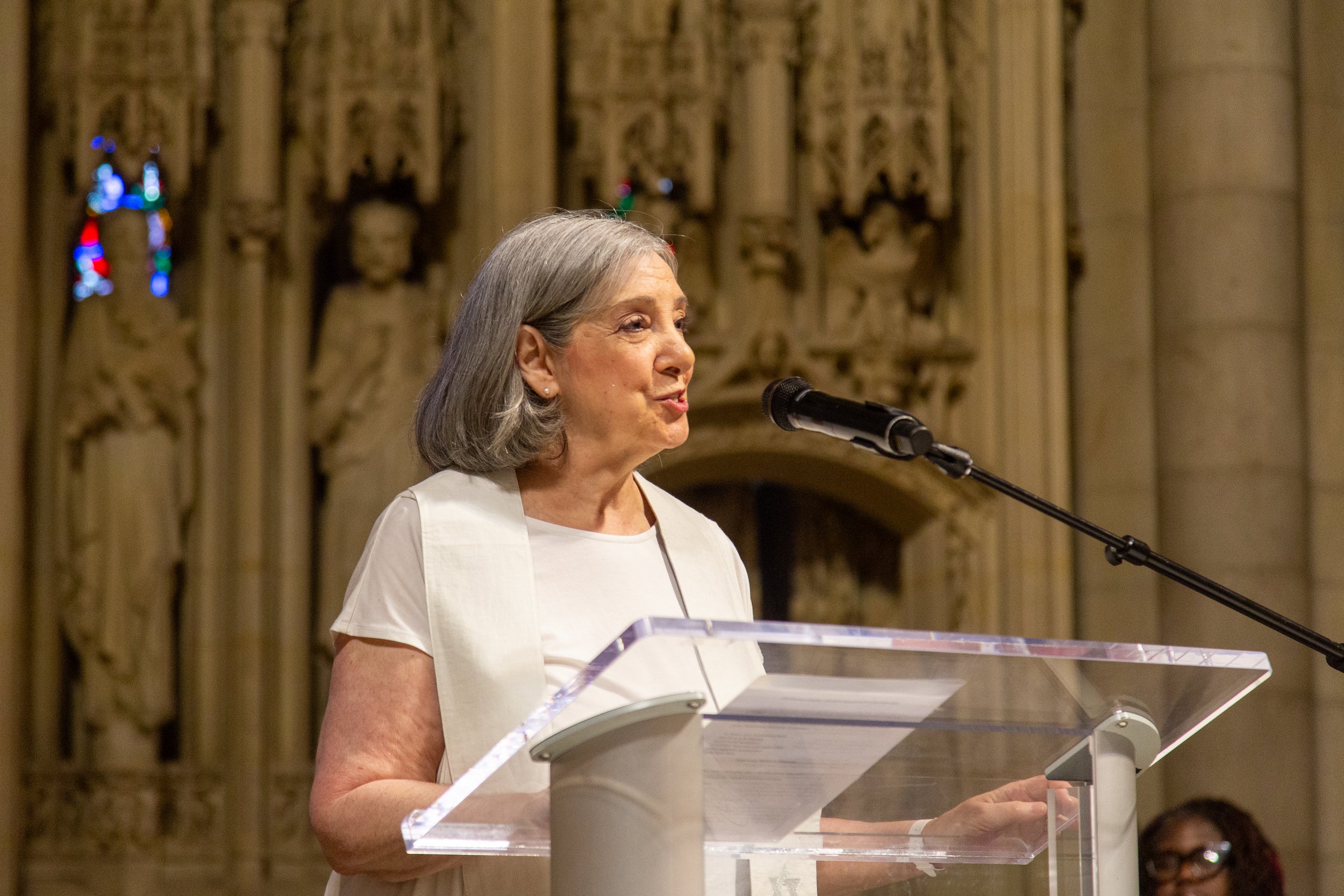 An elderly woman with gray hair speaking at a podium in a church or cathedral.