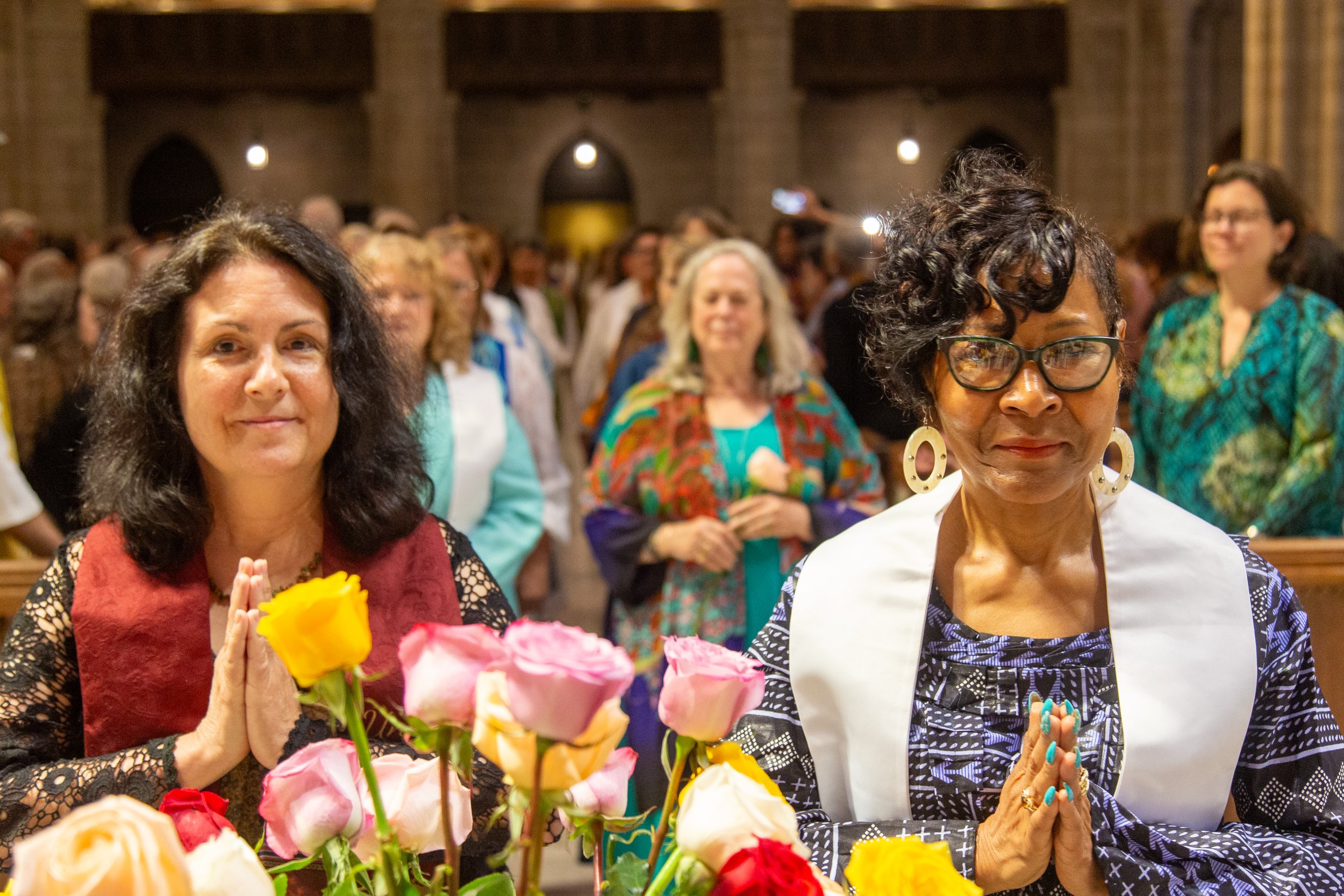 Two women in the foreground with flowers in front of them, one with hands together in prayer and the other with hands together, in a church or cathedral setting with a large group of people in the background.