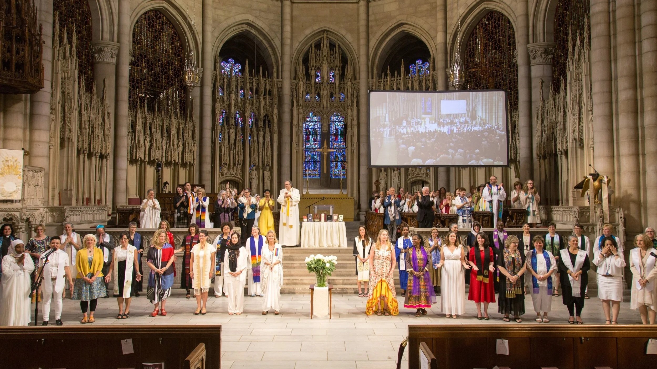 People standing in a church with stained glass windows, some wearing robes and scarves, on a stage during a service or event with a large screen behind displaying the congregation.