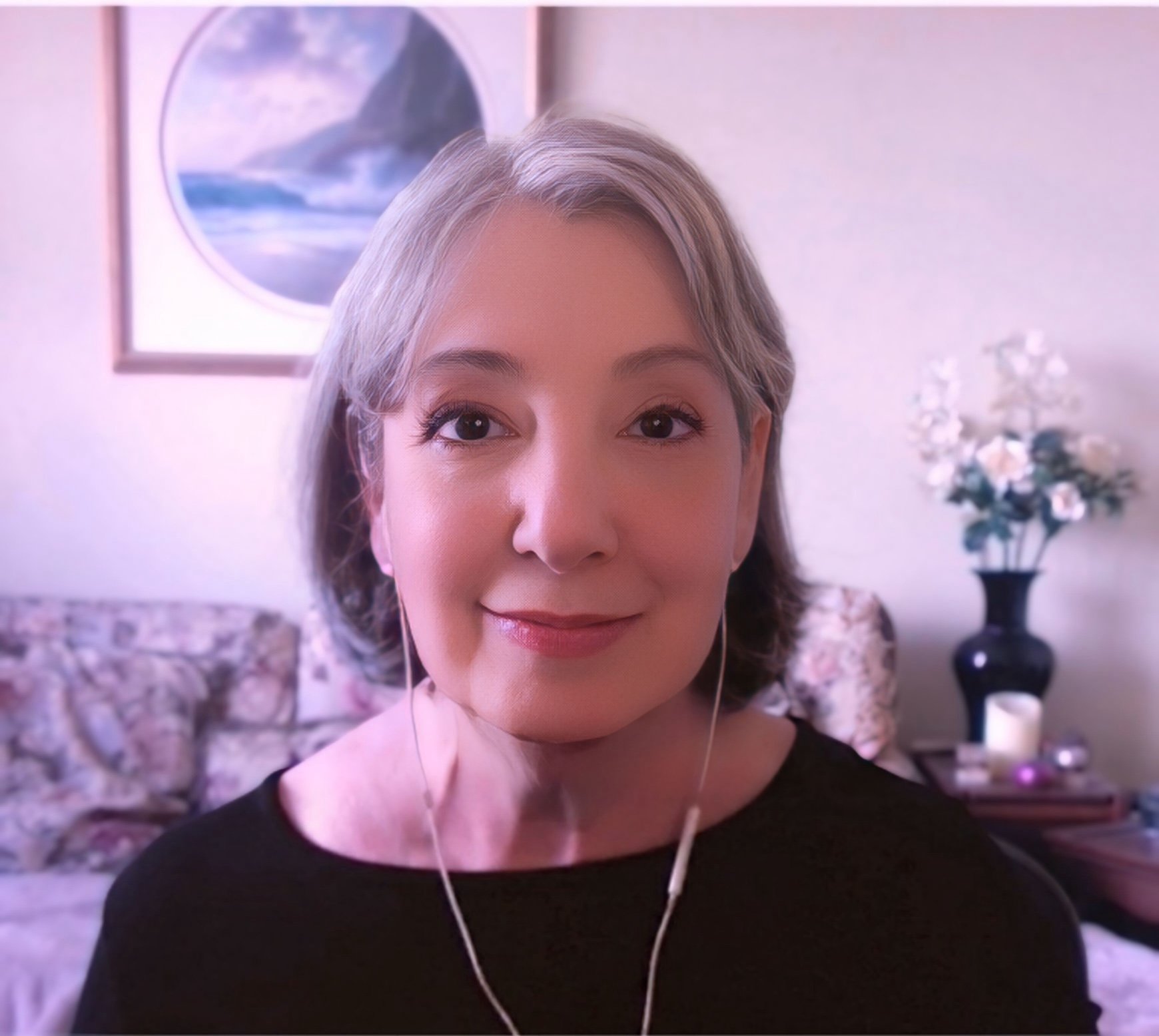 A woman with short gray hair smiling at the camera while wearing earphones in a living room with floral couch, a framed ocean scene painting, and a table with a vase of white flowers and candles.
