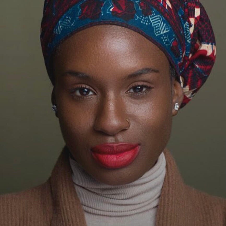 Portrait of a woman with dark skin wearing a colorful headwrap, red lipstick, and a beige turtleneck sweater.
