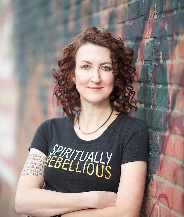 Woman with curly reddish-brown hair smiling, standing against a colorful brick wall, wearing a black T-shirt that says 'Spiritual Rebel-lious' and a tattoo on her left arm.