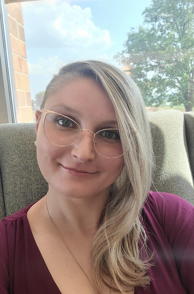 A woman with blonde hair and glasses taking a selfie inside a building near a window with a view of trees and blue sky.