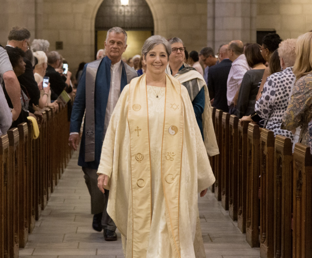 A woman in a religious or ceremonial robe walking down the aisle of a church with a smiling expression, surrounded by an audience taking photos and applauding.