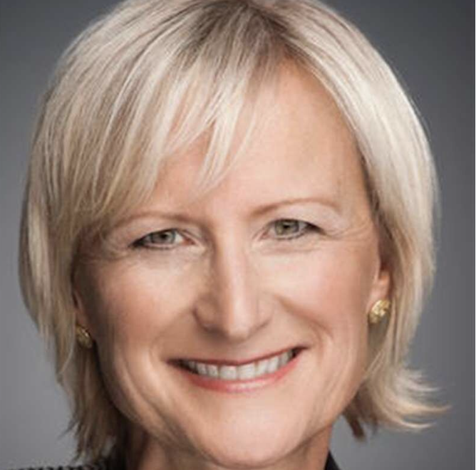 Close-up portrait of a smiling middle-aged woman with short blonde hair and gold earrings against a gray background.