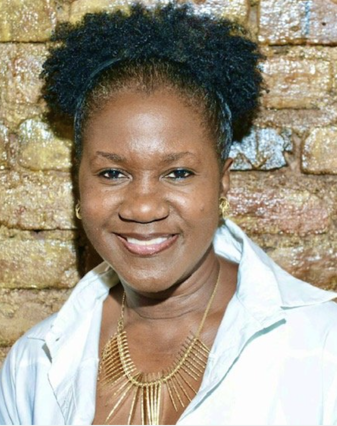 Portrait of a smiling woman with dark, curly hair styled in an updo, wearing a white shirt, gold jewelry, and standing against a brick wall