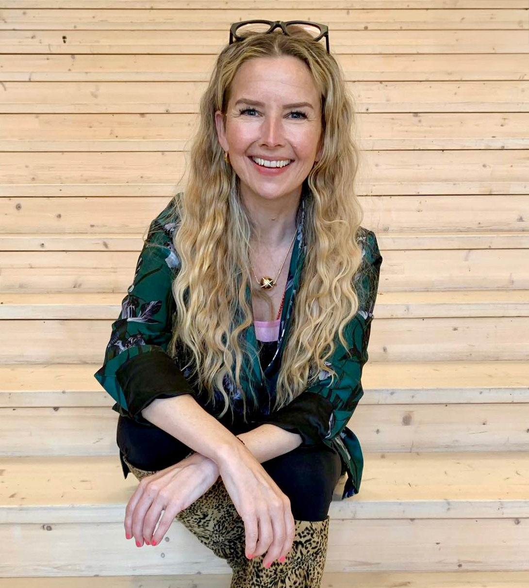 A woman with long, wavy blonde hair, wearing glasses on her head, a patterned black and green blouse, leopard print boots, and jewelry, sitting on wooden stairs against a light wood-paneled background, smiling at the camera.