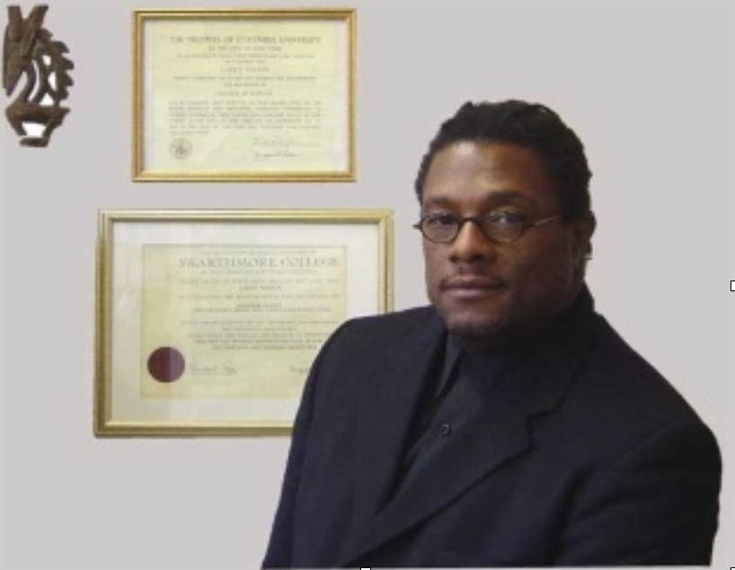 A man with glasses, wearing a dark suit, standing in front of framed diplomas on a white wall.