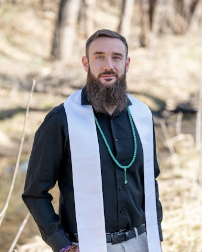 A man with a beard wearing a black shirt, white stole, and turquoise beads outdoors near a stream.