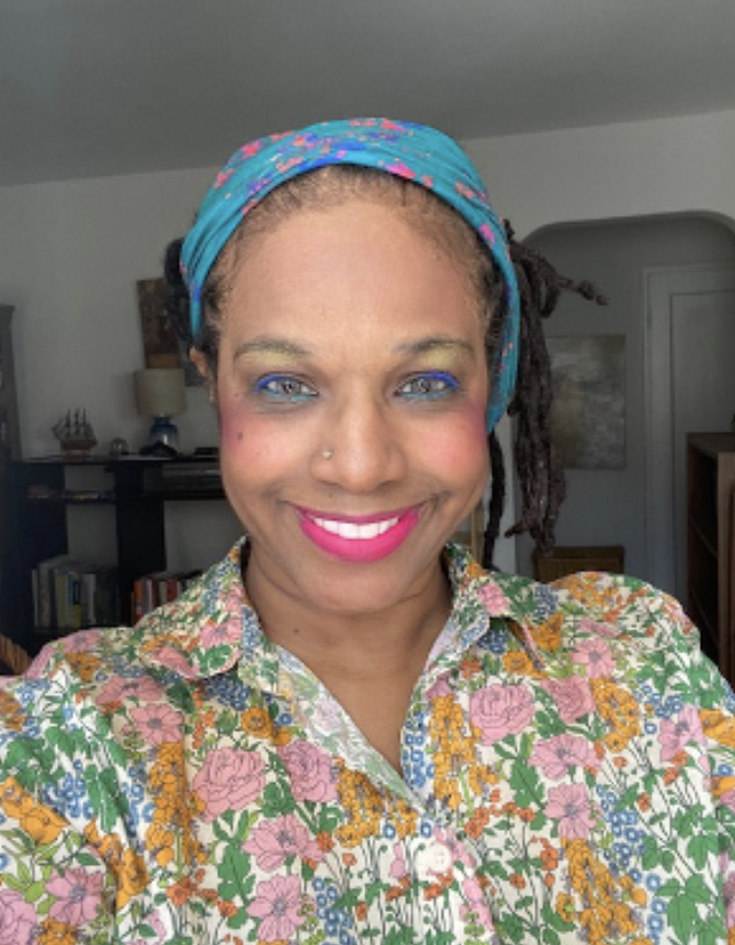 Woman with a bright smile, wearing a colorful floral shirt and a blue headscarf with pink and red patterns, standing in a room with bookshelves and art decor in the background.