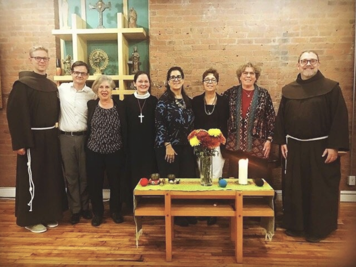 Group of nine people standing inside a room with brick walls, two of whom are wearing brown monk robes. There is a table with flowers and candles in front of them.