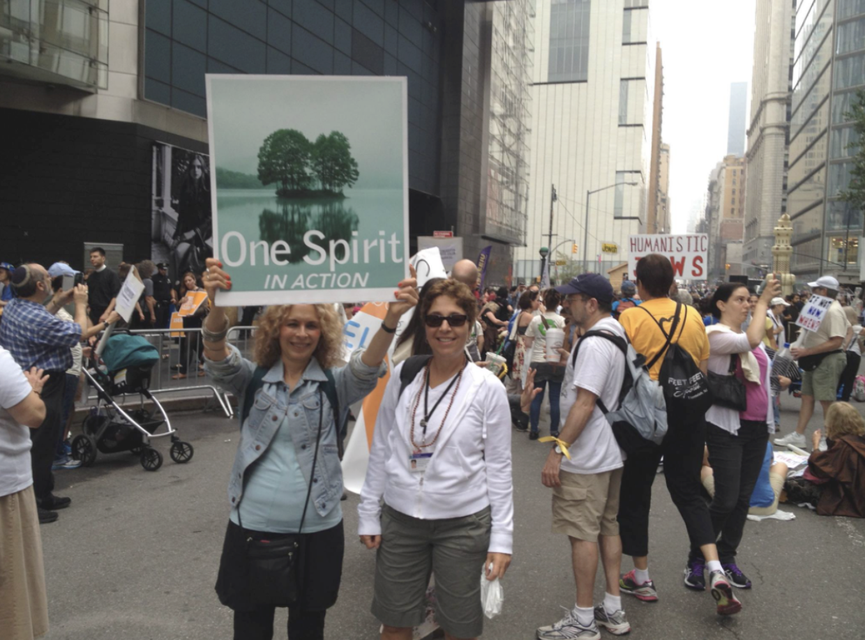 Two women smiling at a protest, with one holding a sign that says 'One Spirit in Action'. Several people around them holding signs, with a city street in the background.