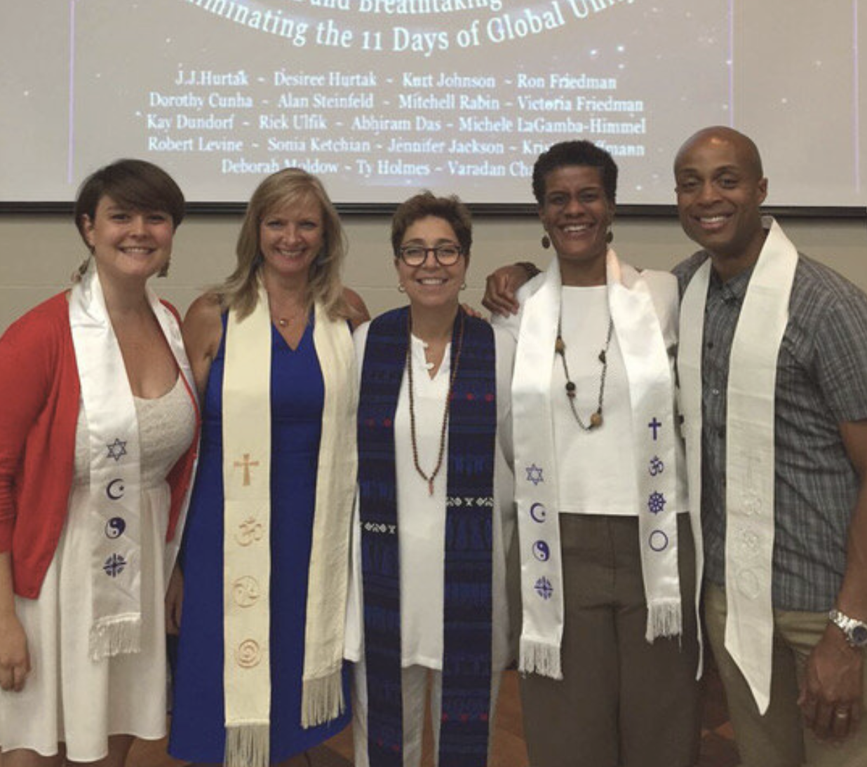 Group of five people smiling and standing together, each wearing a stole with religious symbols, in front of a presentation screen with a list of names and titles.