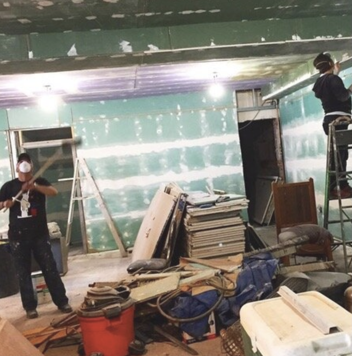 Two workers remodeling a room with green drywall, one on a ladder and the other on a scaffold, surrounded by construction tools, debris, and materials.
