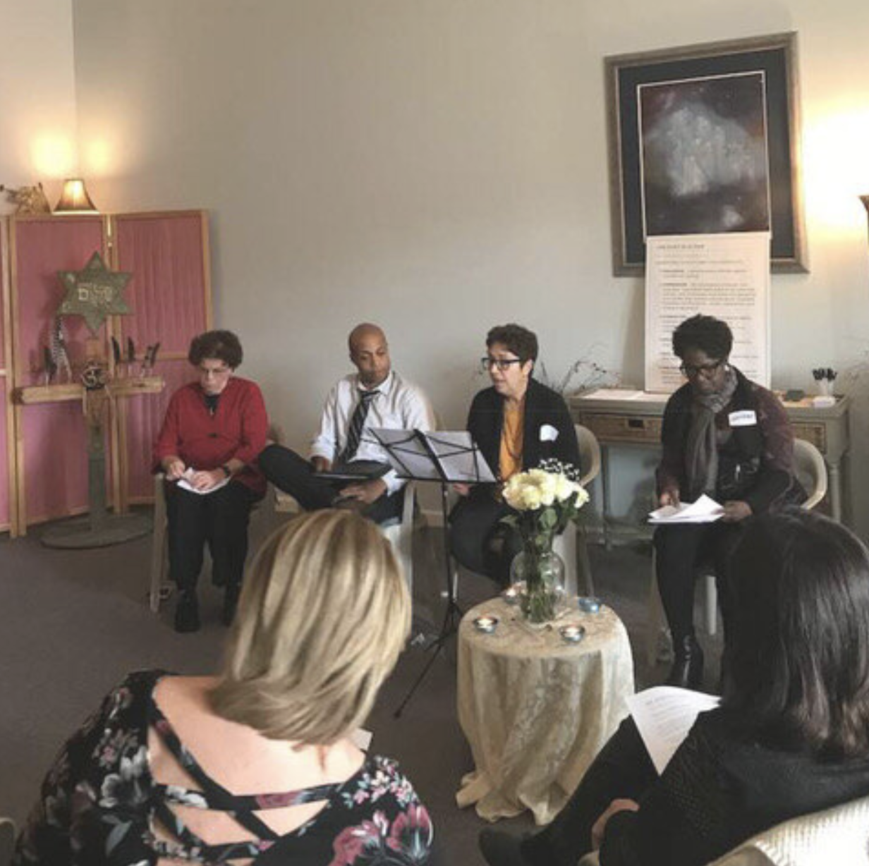 A panel discussion with four people seated, reading from papers or listening, in a decorated room with a pink backdrop, a framed picture of a star, and a vase of flowers on a small table. Audience members are visible in the foreground.