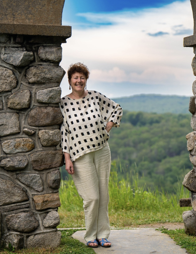 A woman standing between stone arches at a scenic outdoor location, smiling, with a background of rolling green hills and a partly cloudy sky.